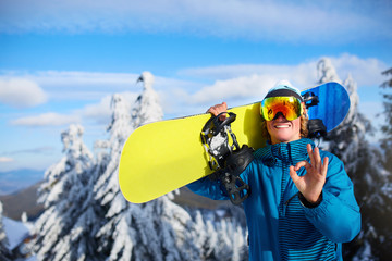 Smiling snowboarder posing carrying snowboard on shoulders at ski resort near forest before...