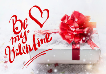 Obraz na płótnie Canvas Be my Valentine text lettering with white gift box, red bow on snow with bokeh and snowfall. Valentines day, declaration of love concept