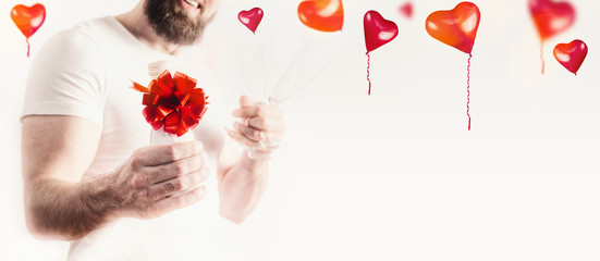 Valentines day background. Young man with bottle of champagne and red bow at white with heart ballons , banner. Male hands holding festive sparkling wine bottle with red ribbon and glasses.