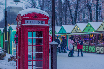 red phone booth and city Christmas fair with people in snowing seasonal winter weather time