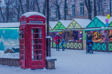 red phone booth and city Christmas fair with people in snowing seasonal winter weather time