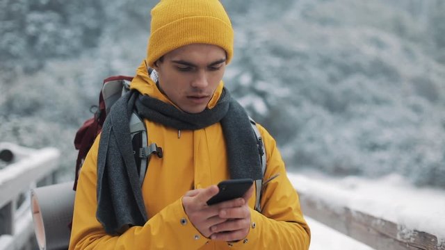 A young tourist standing on a bridge in the snowy mountains with a smartphone in his hands. Communication, social networks, travel concept