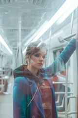 Young beautiful girl with brown hairs and bright make up, dressed in blue coat, black hoodie, sneakers and checkered trousers posing in metro carriage. Posing behind the glass