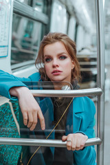 Young beautiful girl with brown hairs and bright make up, dressed in blue coat, black hoodie, sneakers and checkered trousers posing in metro carriage. Posing behind rails and glass