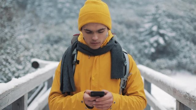 A young tourist walks on a bridge in the snowy mountains with a smartphone in his hands. Communication, social networks, travel concept