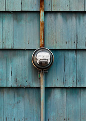 Electricity line drop residential dial power meter mounted on exterior wooden shingle siding wall with faded blue paint