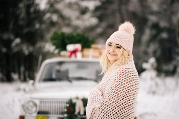 The girl is preparing for Christmas, is covered with a blanket on the background of a retro car, the roof of which is a Christmas tree, gifts and a wreath in the winter snowy forest.
