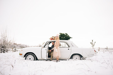 A guy and a girl ride in a retro car decorated with a Christmas tree and presents in a snowy forest. The concept of a winter Christmas love story