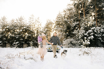 Fototapeta na wymiar A happy young family with a small child is preparing for Christmas, walking with a husky dog on the background of a retro car, on the roof of a Christmas tree and gifts in the winter snowy forest.