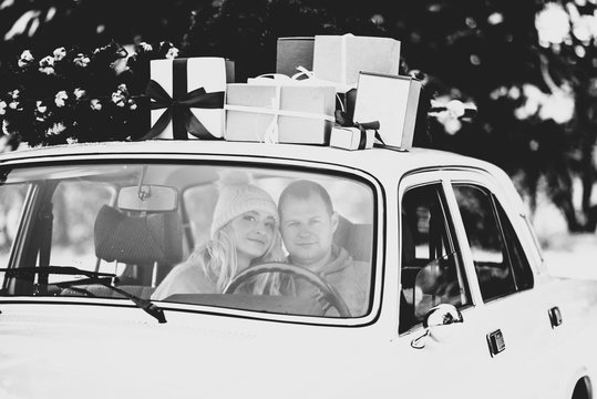 A guy and a girl ride in a retro car decorated with a Christmas tree and presents in a snowy forest. Black and white photography