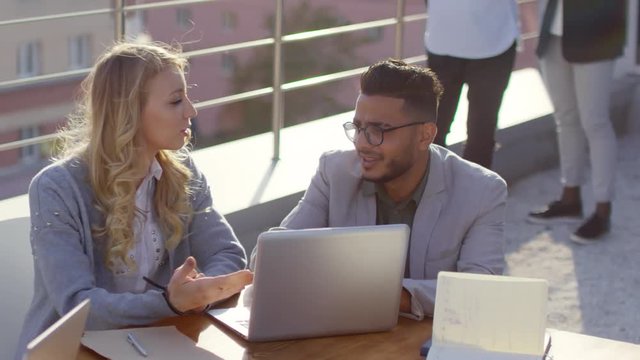Tilt down of young middle eastern man and Caucasian blonde woman sitting at table on rooftop, smiling and discussing business project on laptop
