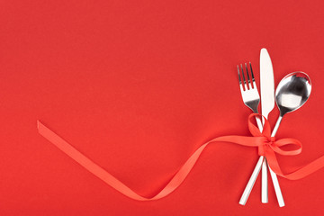 view from above of fork, knife and spoon wrapped by red festive ribbon isolated on red, st valentine day concept