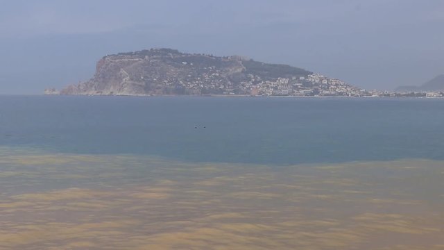 View on Bedesten Alanya from beach. View from dirty to clear water. Island in the sea. Overcast weather.