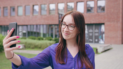 Fototapeta premium A happy smiling brunette woman with glasses using a phone and taking a selfie in the city street. Close-up shot. Soft focus