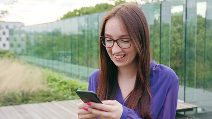 A happy smiling brunette woman with glasses using a phone in the city street. Close-up shot. Soft focus