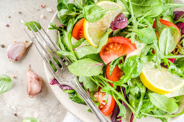 Fresh spring detox mix salad with vegetables (cucumber, lemon, tomato, arugula, and baby spinach), on light slate, stone or concrete background. Top view copy space