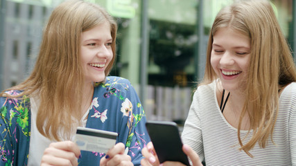 Attractive young ladies using a phone and holding a credit card in town. Medium shot. Soft focus