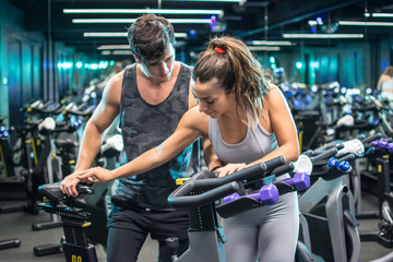 Young attractive woman with handsome trainer setting up exercise bike before cycling workout in gym 