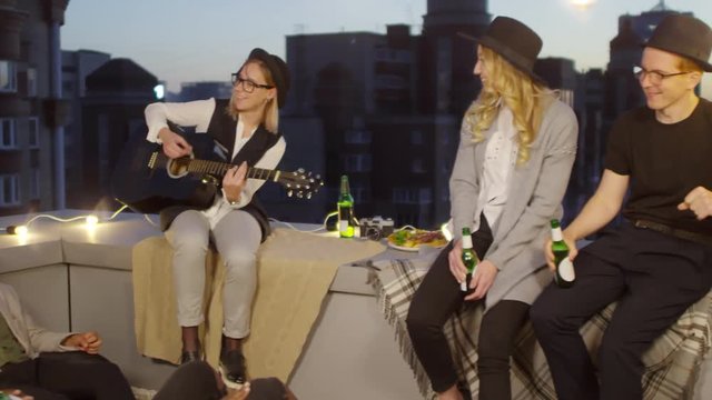 Multiracial hipster friends sitting on blankets and bean bags, holding beer bottles up and enjoying music while young woman playing the guitar at summer party on urban rooftop decorated with lights