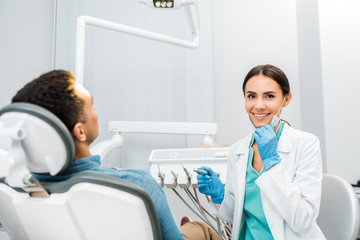 cheerful dentist holding drill and smiling near african american patient