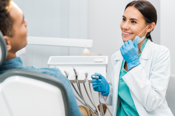 cheerful female dentist holding mask and drill and smiling near patient - Powered by Adobe