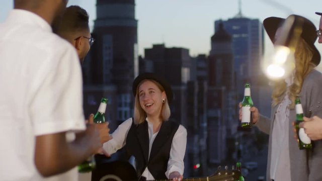 Multi ethnic group of young happy friends laughing and dancing with beer bottles while woman playing the guitar at summer party on urban rooftop in the evening