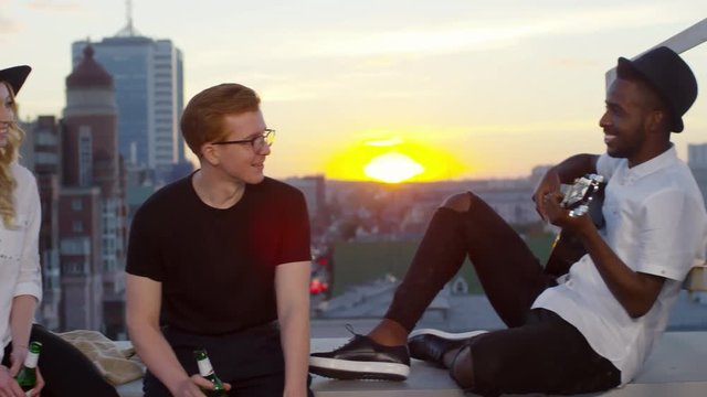 Young african american man playing the guitar and smiling while his friends sitting with beer bottles and enjoying the music on urban rooftop at sunset