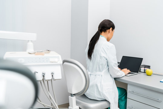 Back View Of Female Doctor In White Coat Typing On Laptop