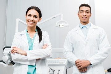 cheerful  female doctor standing near african american coworker in white coat with crossed arms