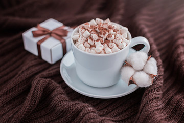 Coffee cup with marshmallow and gift box with brown ribbon on warm knitted brown blanket.
