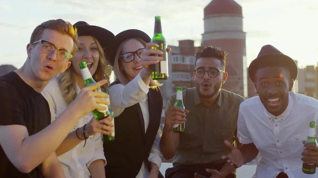 Multiethnic group of young hipster friends smiling, yelling and raising beer bottles to camera at summer party on rooftop terrace