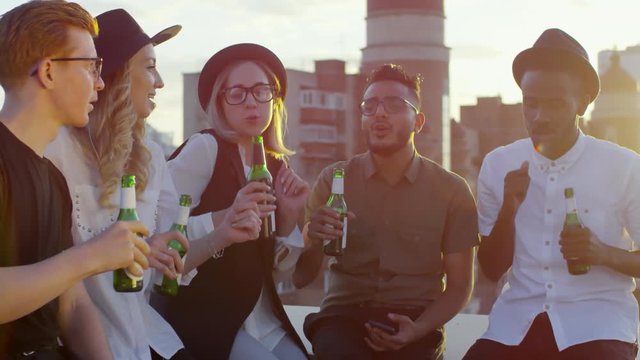 Young hipster friends of diverse ethnicities smiling and dancing with beer bottles at summer party on rooftop terrace at sunset