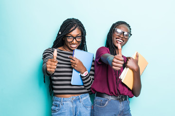Young afro american student girls show thumbs up and holding netobooks isolated on pastel blue background