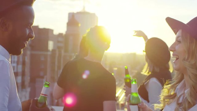 Young pretty woman and african american man in fedora hats holding beer bottles, laughing and dancing together at party with friends on urban rooftop at sunset