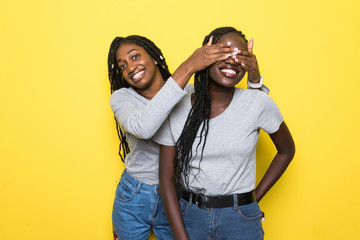 Portrait of two young african women make surprise and cover eyes isolated on yellow background