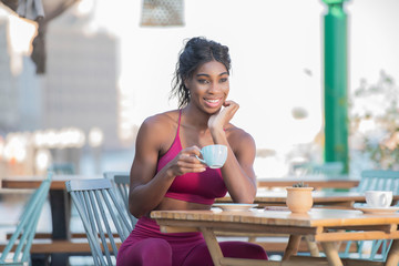  Beautiful tall athletic African American Woman wearing a bright pink workout outfit sits in an outdoor cafe drinking coffee or tea in deep though on a bright sunny day