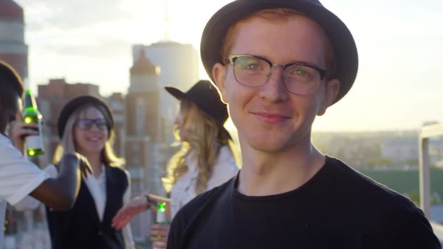 Portrait of young redhead man wearing glasses and fedora hat looking at camera and smiling while his multiracial friends dancing with beers in the background at party on city rooftop at sunset