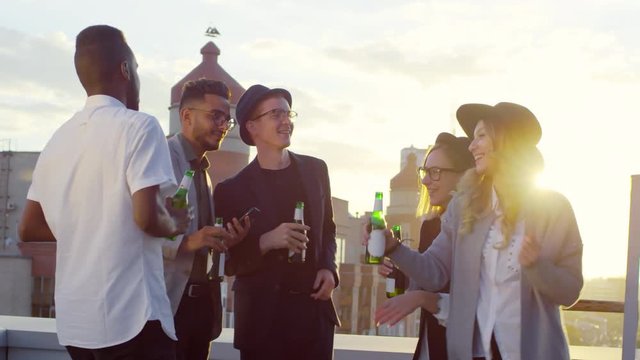 Multiracial group of young hipster friends dancing and drinking beer from bottles at outdoor party on urban rooftop at sunset