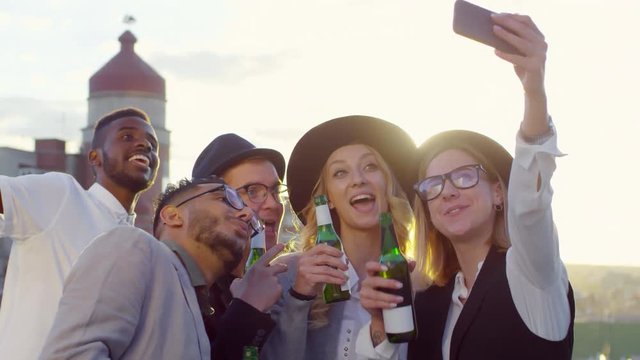 Group of young diverse friends holding beer bottles, smiling and posing for smartphone camera while taking selfie together at party on rooftop