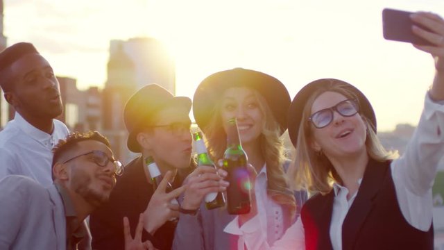 Group of young happy multiethnic friends holding beer bottles, smiling and posing for smartphone camera while taking selfie together on rooftop at sunset