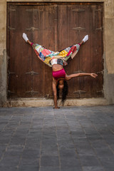Obraz na płótnie Canvas Beautiful tall athletic African American Woman does a one handed handstand pose wearing a bright pink sports bra and a multicolored pant against an old wooden door
