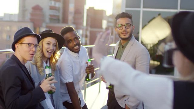 Multiethnic young friends standing on rooftop at sunset, holding beer bottles, smiling and waving at camera while female photographer taking picture of them