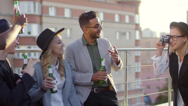 Young multiethnic friends smiling and holding beer bottles while posing for camera during rooftop party; female photographer in fedora hat taking picture of them