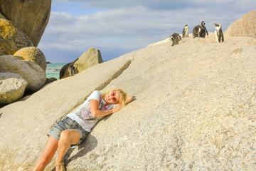 Fototapeta na wymiar Happy blonde tourist woman resting on the rocks of Boulder Beach in summer, a popular travel destination of a colony of African penguins. Cape Peninsula near Cape Town in Western Cape, South Africa.