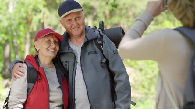 Tilt up shot of elderly woman with backpack taking photo of happy senior couple while hiking in forest