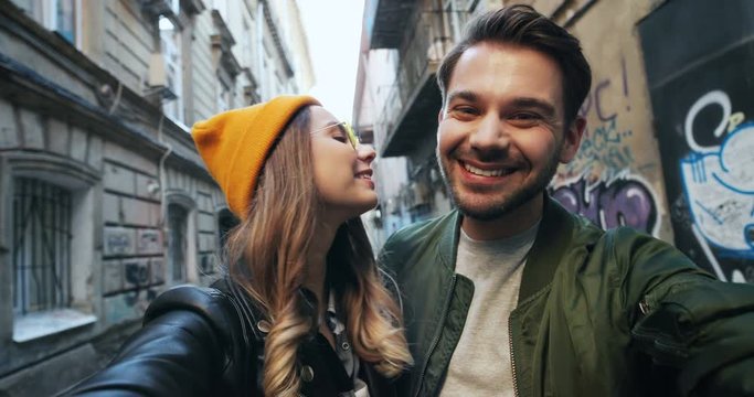 Joyful young happy Caucasian couple in stylish looks smiling and posing to the camera while taking selfie picture at the street. POV. Close up. Outdoor.