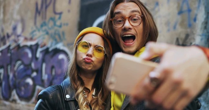 Close up of the Caucasian happy young boyfriend and girlfriend making funny faces while taking selfie photos on the smartphone camera at the graffity wall. Portrait. Outdoor.