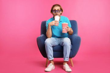 Emotional man with 3D glasses, popcorn and beverage sitting in armchair during cinema show on color...