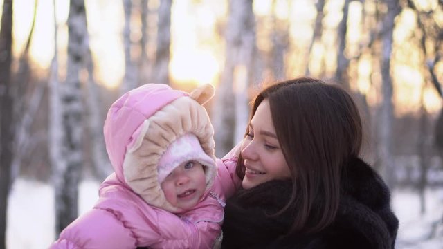 Close-up of young happy mother with cheerful child in winter outdoors. Baby girl in pink jumpsuit smiling in mom's hands on walk in park winter