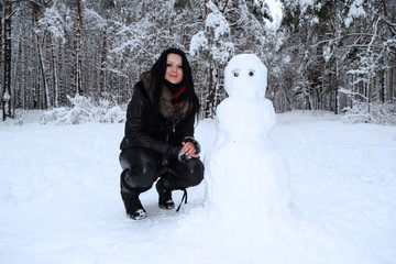 Fototapeta na wymiar Young girl in black clothes sitting near white snowman in winter forest.